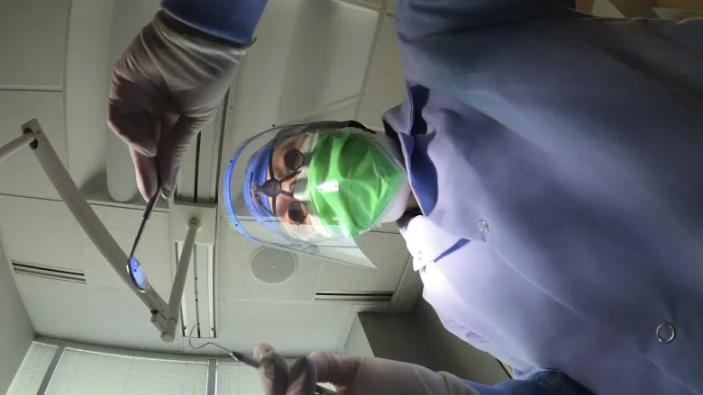 A dentist holds instruments looking down at the camera. They are wearing a face shield and a mask.